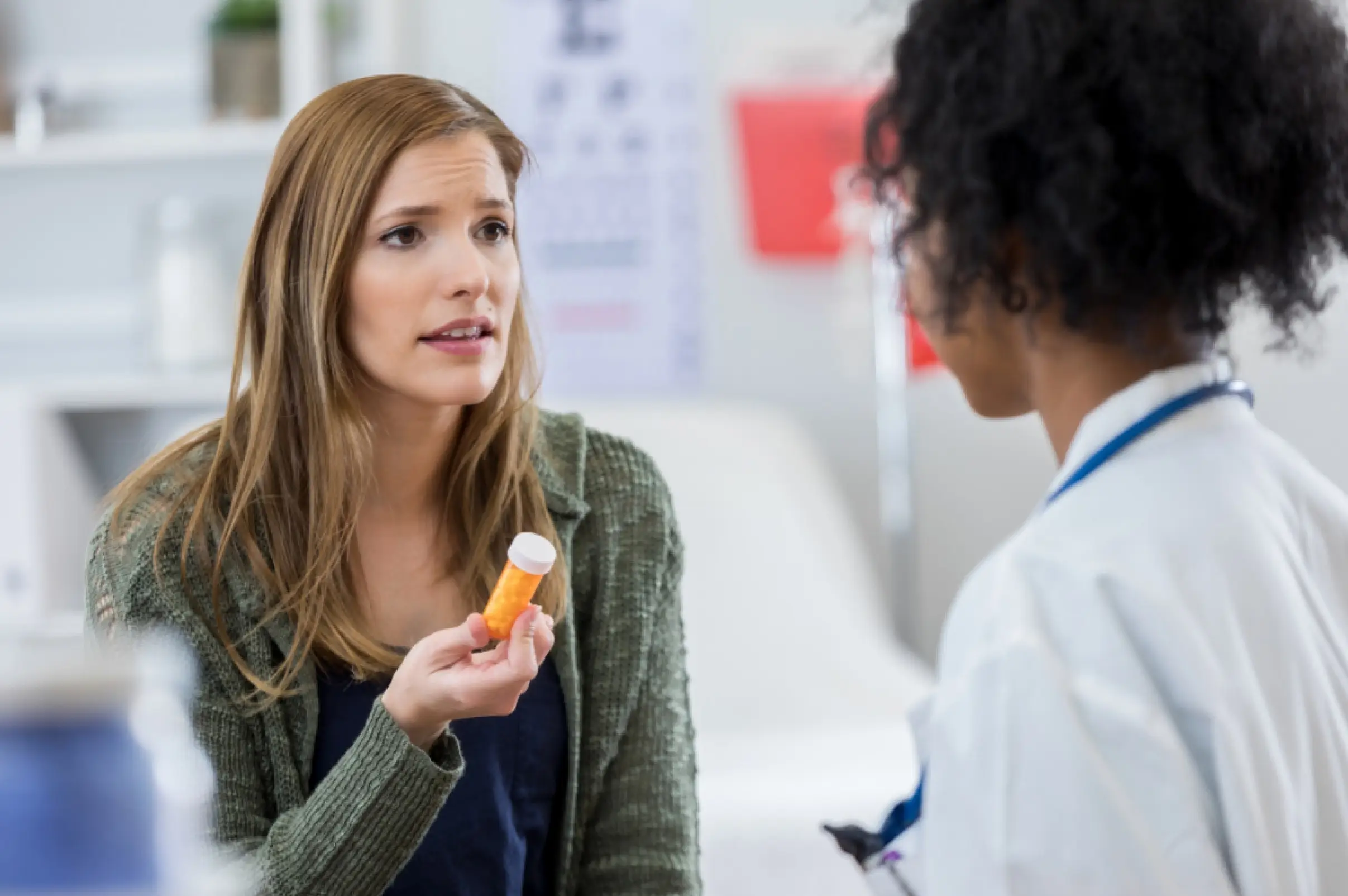 Woman holding a prescription pill bottle while speaking with a healthcare provider in a medical office.